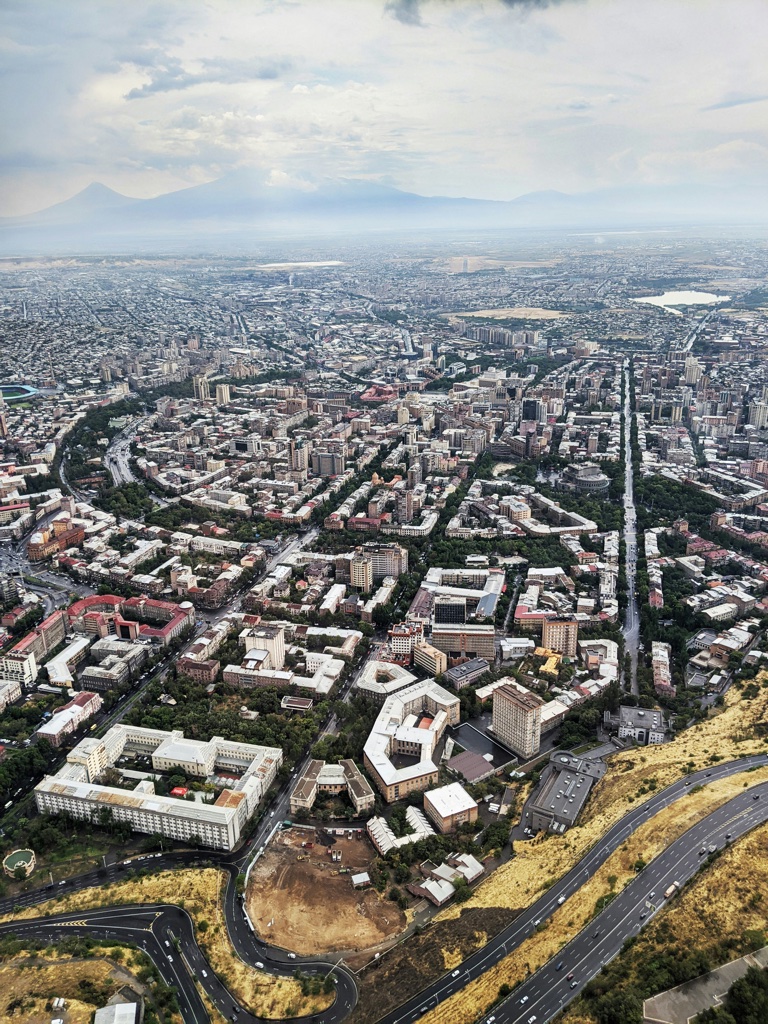 Yerevan from the air, dense pink and grey blocks packed around a tree lined avenue running to the horizon, a dry yellow hillside and a curving highway across the foreground, and mountains faint behind the haze.