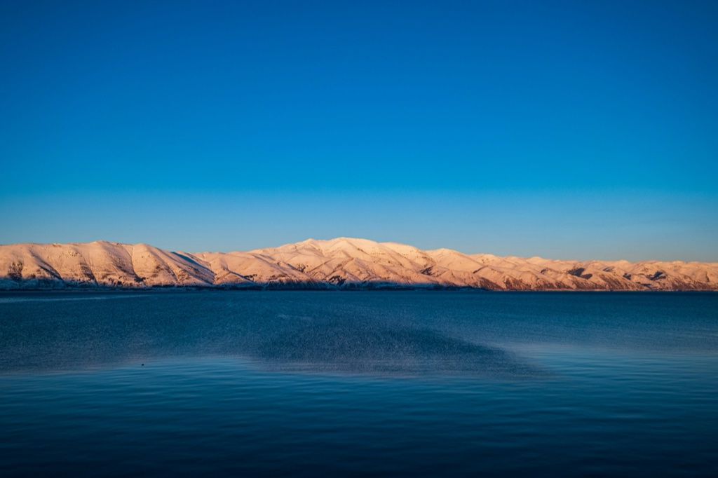 A long low ridge of snow covered hills lit orange by a low sun, seen across deep blue open water under a cloudless dark blue sky.