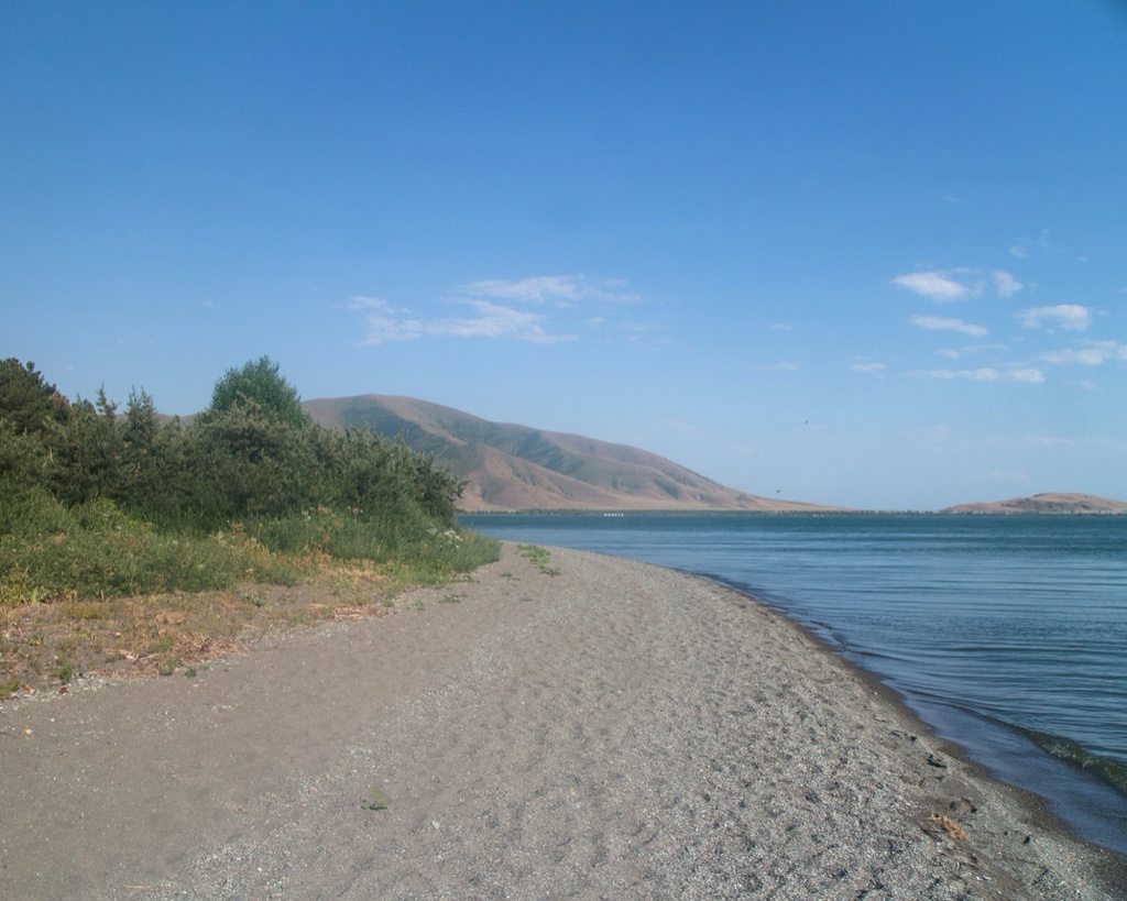 A grey pebble beach curves away along a still lake, scrub and small trees crowding the bank on the left and a bare brown ridge running along the far shore.