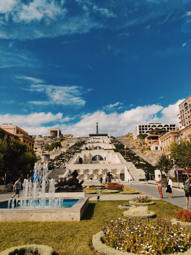 A wide flight of stone steps climbing a hillside in Yerevan above a fountain and beds of flowers, with people walking past in the sun.