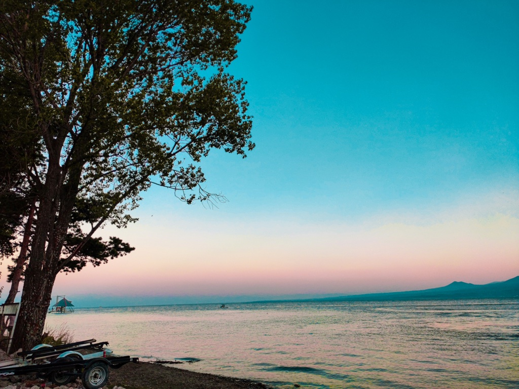 A lake at dusk under a teal and pink sky, a tall tree silhouetted on the left bank, a boat trailer parked on the shingle below it and a headland low on the horizon.