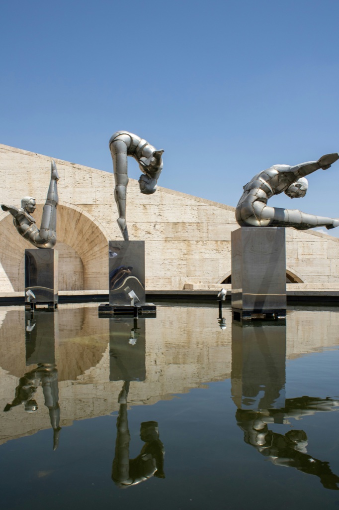 Three steel diver sculptures on stone plinths in front of a long arcaded wall of pale cut stone, mirrored in the still pool beneath them.