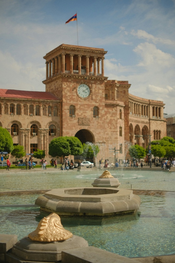 A clock tower and arcades built of pink and orange volcanic stone stand above a fountain, with the Armenian flag flying from the roof and people crossing the square below.